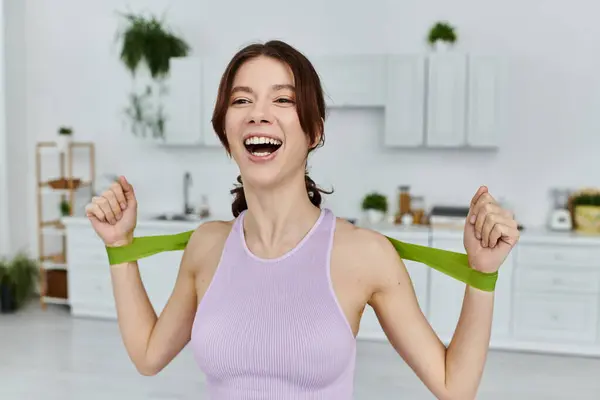 A young woman in sportswear laughs as she stretches with a resistance band in a modern apartment. — Stock Photo