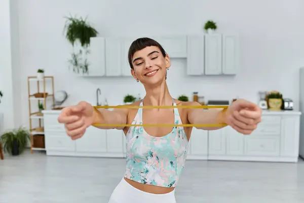 A woman smiles as she stretches with a yellow resistance band in a modern apartment. — Stock Photo