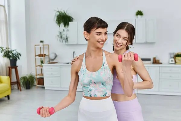 Two women, in sportswear, smile and work out with pink dumbbells together in a modern apartment. — Stock Photo
