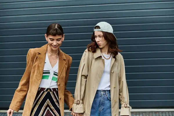 Two stylish women dressed in trendy outfits, walk hand in hand against a contemporary backdrop. — Stock Photo
