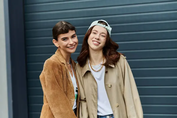 Duas jovens mulheres em roupas da moda, sorrindo para a câmera na frente de um pano de fundo da cidade. — Fotografia de Stock