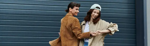 Two trendy women, one in a brown jacket and the other in a beige jacket and cap, laugh and stroll in front of a blue garage door — Stock Photo