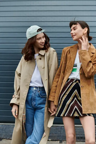 A young lesbian couple, dressed in trendy outfits, walk hand-in-hand in front of a gray garage door. — Stock Photo