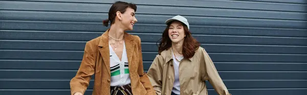 Two young women in trendy outfits walk side by side, laughing together against a blue wall. — Stock Photo