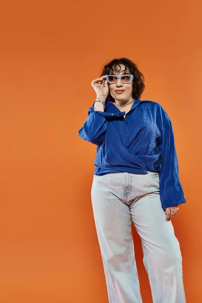 A woman in a blue shirt and light-wash jeans confidently adjusts her sunglasses. — Stock Photo