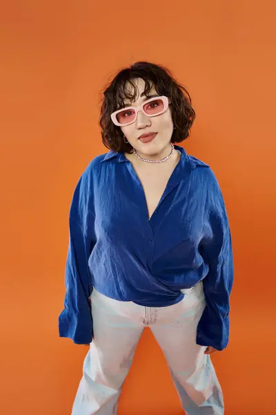 A woman poses in a blue shirt and sunglasses against an orange backdrop. — Stock Photo
