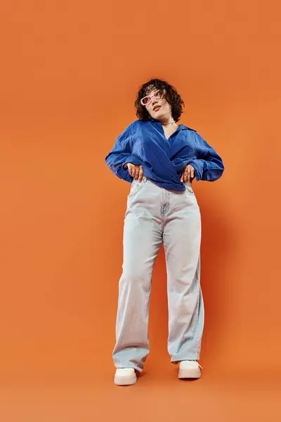 Una mujer con una camisa azul se levanta contra una pared naranja, manos en las caderas. - foto de stock