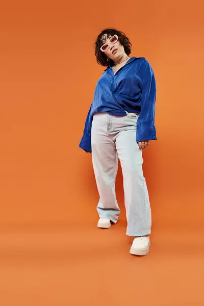 A young woman in a blue shirt and sunglasses stands confidently on an orange backdrop. — Stock Photo