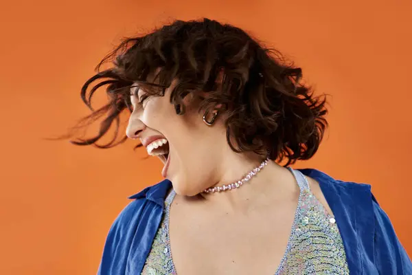 Happy woman in blue shirt and sparkly top shouts joyfully against orange backdrop — Stock Photo