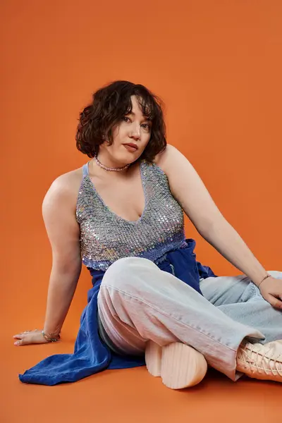 Frau in Paillettenoberteil und Jeans sitzt auf orangefarbenem Hintergrund und blickt von der Kamera weg — Stockfoto