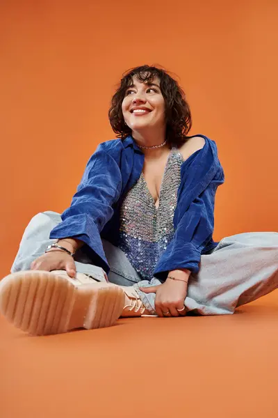 Woman in blue shirt and sparkly top sits against orange backdrop, looking joyful — Stock Photo