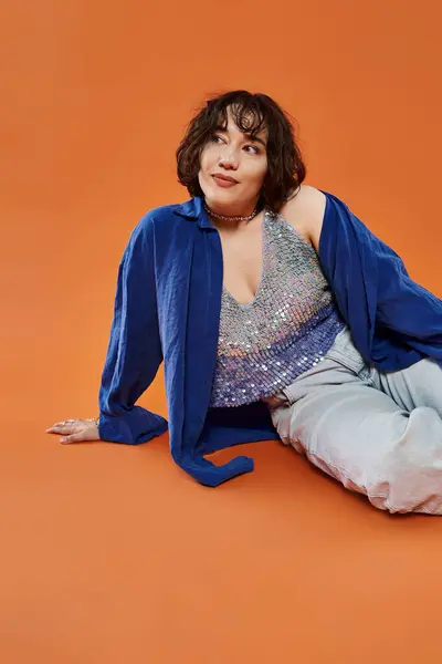 Una mujer se sienta sobre un fondo naranja, vistiendo una camisa azul y una blusa brillante. - foto de stock