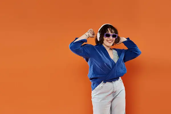 Woman in blue shirt and white jeans dances with sunglasses and headphones on orange background — Stock Photo