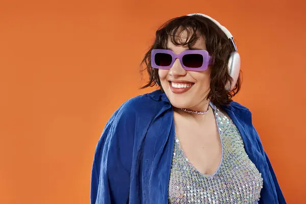 A woman wearing purple sunglasses and a blue jacket smiles against an orange background. — Stock Photo