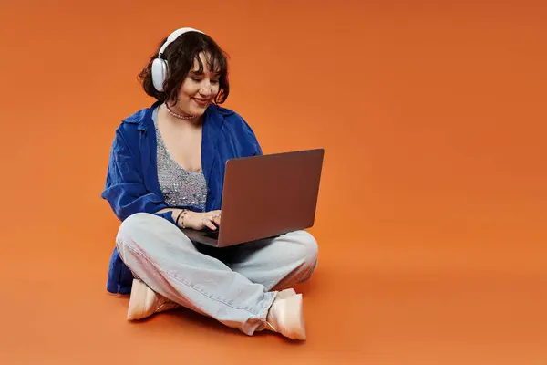 Una mujer con auriculares se sienta en el suelo, usando una computadora portátil. - foto de stock