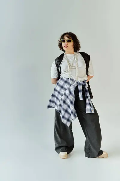 Una mujer con una camisa a cuadros y pantalones negros está de pie con confianza en un estudio blanco. - foto de stock