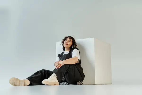 A woman in black pants and a white shirt sits against a white cube, eyes closed, radiating serenity. — Stock Photo