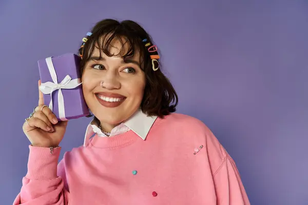A woman smiles brightly, holding a purple gift box to her ear as if listening to its contents. — Stock Photo