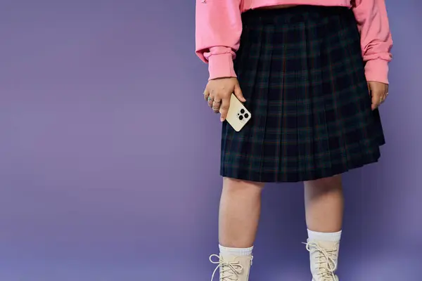 Une femme en jupe à carreaux et pull rose tient un téléphone sur fond de lavande. — Photo de stock