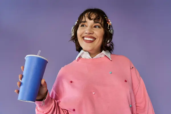 A woman in a pink sweatshirt and headphones holds a blue cup with a straw, smiling brightly. — Stock Photo
