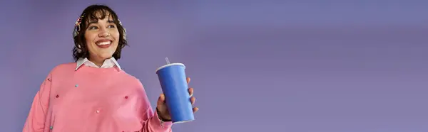 A smiling woman in a pink sweater holds a blue cup against a purple background. — Stock Photo