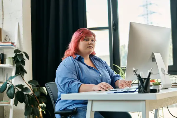 A plus size woman sits at a desk, typing on a computer, with a determined expression. — Stock Photo