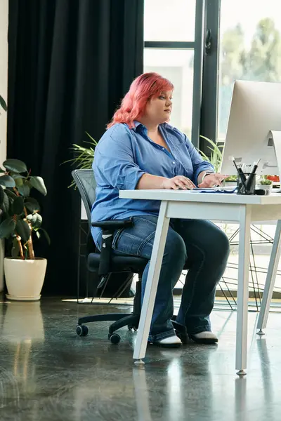 Женщина plus size с ярко-розовыми волосами сидит за письменным столом, работая за компьютером. — стоковое фото