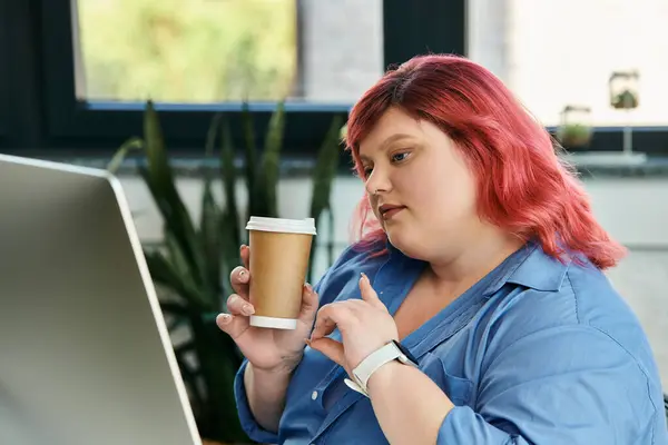 Una donna d'affari plus size in una camicia blu tiene una tazza di caffè e guarda uno schermo del computer. — Foto stock