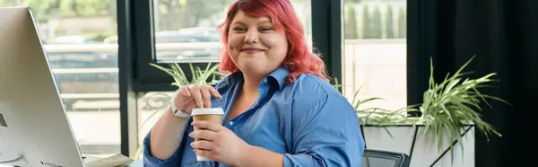 A plus size businesswoman with pink hair sips coffee while sitting at a desk in a modern office. — Stock Photo