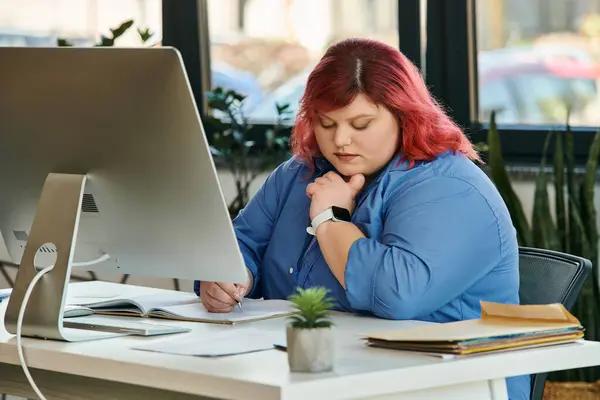 Une femme d'affaires de taille plus avec les cheveux roux écrit à un bureau, regardant un ordinateur — Photo de stock