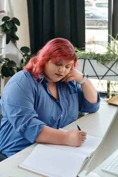 A plus size businesswoman with pink hair sits at a desk, writing in a notebook. — Stock Photo