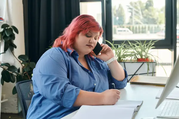 Une femme d'affaires de taille plus avec les cheveux roses prend des notes lors d'un appel téléphonique dans un espace de bureau lumineux. — Photo de stock