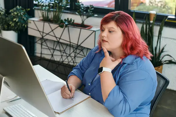 A plus size businesswoman thoughtfully takes notes while looking at a computer screen. — Stock Photo