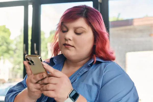 Una donna plus size con i capelli rosa usa il suo telefono, seduta davanti a una finestra. — Foto stock