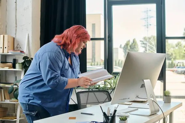 A plus size woman stands by her desk, engrossed in a book while a computer screen waits nearby. — Stock Photo