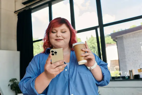 Eine Plus-Size-Geschäftsfrau mit roten Haaren lächelt, als sie ihr Handy kontrolliert, während sie eine Tasse Kaffee in der Hand hält.. — Stockfoto