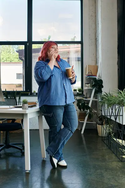 Una mujer de negocios de tamaño grande en azul se apoya sobre un escritorio, disfrutando del café en una oficina moderna. - foto de stock