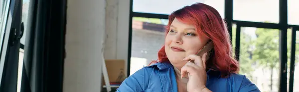 A plus size businesswoman with vibrant red hair smiles as she speaks on her phone, sitting by a large window. — Stock Photo