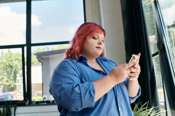 Une femme d'affaires de taille plus avec les cheveux roses utilise son téléphone près d'une grande fenêtre. — Photo de stock