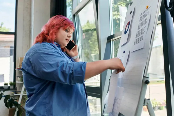 A plus size woman on a phone call stands near a whiteboard, reviewing project notes. — Stock Photo