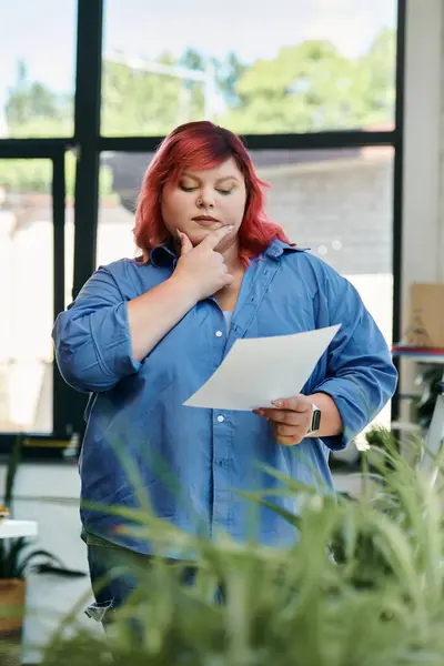 Uma mulher plus size com cabelo vermelho vibrante revisa cuidadosamente um documento. — Fotografia de Stock