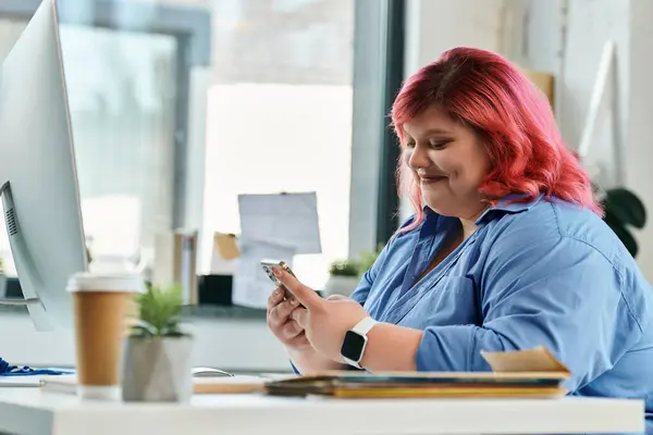 A plus size businesswoman with pink hair sits at her desk, smiling as she uses her smartphone. — Stock Photo