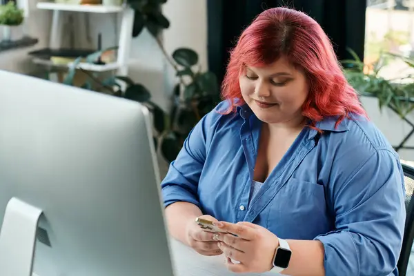 A plus size businesswoman with pink hair checks her phone while sitting in front of a computer. — Stock Photo