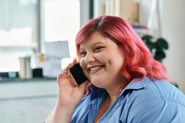 Una donna d'affari plus size con i capelli rosa, indossando una camicia blu, parla al telefono con un sorriso. — Foto stock