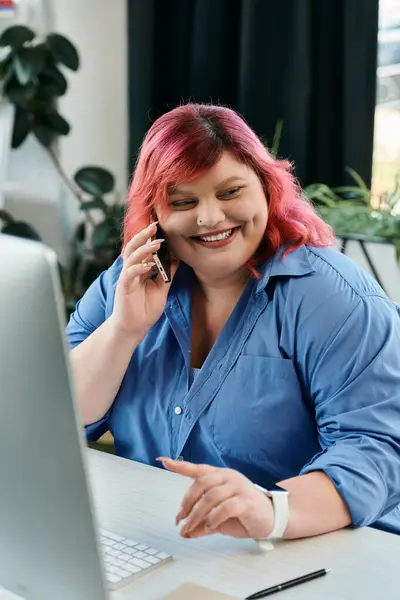 A plus size businesswoman with pink hair talks on the phone while sitting at her desk, smiling. — Stock Photo
