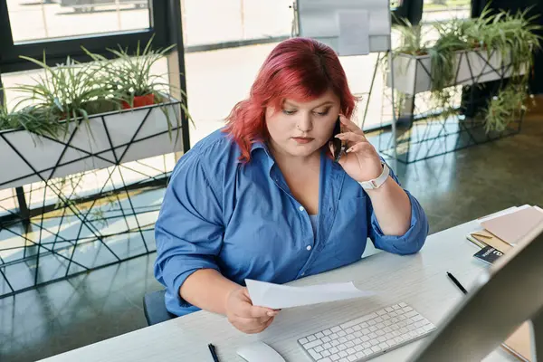 Женщина plus size в голубой рубашке берет телефонный звонок, рассматривая документ на своем рабочем столе. — стоковое фото