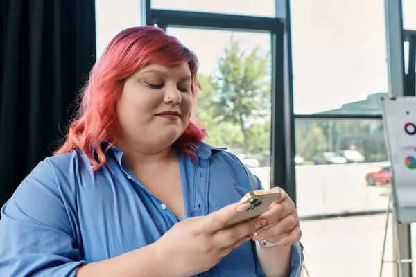 A plus size woman with pink hair and a blue shirt checks her phone in front of a window. — Stock Photo