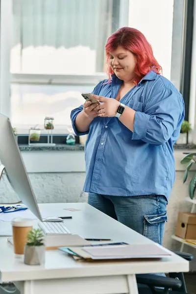 Eine Plus-Size-Frau im blauen Hemd checkt ihr Handy, während sie neben einem Schreibtisch steht. — Stockfoto