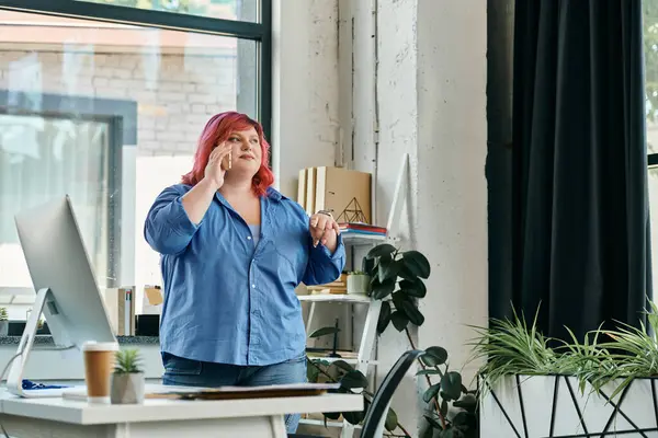 A plus size woman with vibrant pink hair takes a phone call while standing at a desk in a modern office. — Stock Photo