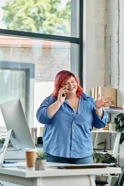 A plus size woman with pink hair smiles while talking on a phone in an office. — Stock Photo